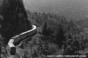 Doubledeck passenger train climbs in s-curves, along the precipice towards a tree-covered valley.