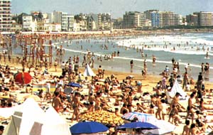 Crowded beach, with a town behind.