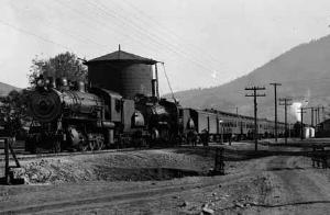 Doubleheaded steam train in a rural station between mountains.